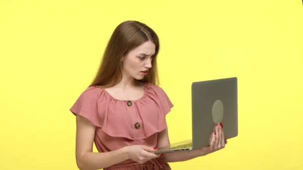 迷人的棕色头发的女孩穿着雅致的衣服在笔记本电脑上工作 得到了新闻 感到惊喜 高兴得紧紧抓住拳头 在黄色背景下隔离的室内拍摄 — 图库视频影像