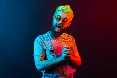 Tişörtlü ve kot pantolonlu çocuksu hippi dili dışarı çıkmış lolipop yalıyor, aç arzuluyor, tatlı şekerlemelere bayılıyor. Renkli neon ışık, kapalı stüdyo çekimi..