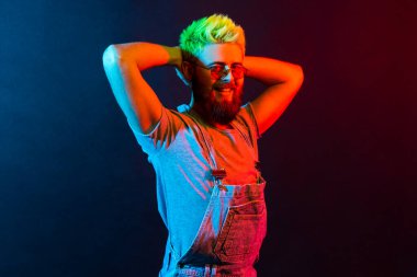 Yüzünde olumlu ifade olan çekici bir genç adam kameraya büyüleyici bir gülümsemeyle bakıyor, el kaldırıyor, tulum giyiyor. Renkli neon ışık, kapalı stüdyo çekimi..