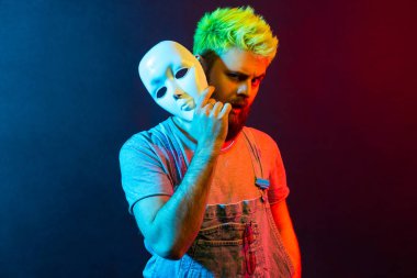 Beyaz maskeli şık saç kesimi olan yakışıklı hippi portresi kaşlarını kaldırmış, şaşkın bakışlı, çoklu kişilik bozukluğu olan. Renkli neon ışık, kapalı stüdyo çekimi..