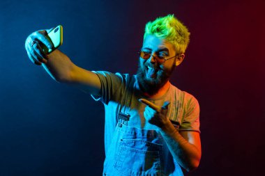 Handsome hipster bearded man wearing t shirt and denim overalls, broadcasting live stream, points to camera with finger, grimacing, winking and showing tongue. Colorful neon light, indoor studio shot clipart