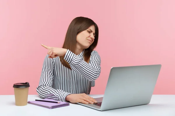 Irritated bossy business woman sitting at workplace and pointing finger aside showing exit, asking to leave office, firing from work. Indoor studio shot isolated on pink background