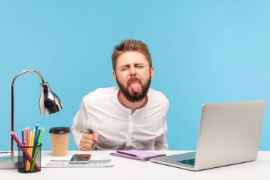 Masasında dizüstü bilgisayarla ofis işyerinde oturmuş, kameraya yaramaz itaatsiz bir ifadeyle bakıyor. Mavi arka planda kapalı stüdyo çekimi