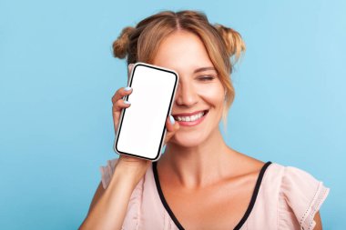 Yazlık elbiseli, gülümseyen ve yüzünün yarısını cep telefonuyla kapatan neşeli sarışın kadın portresi neşeli ve kaygısız görünüyor. Mavi arka planda kapalı stüdyo çekimi.