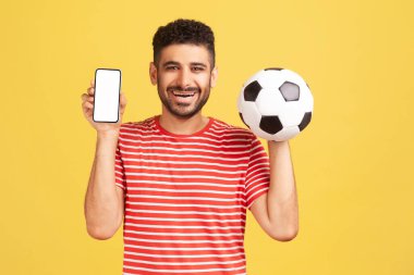 Çizgili tişörtlü, sakallı, beyaz gösterişli akıllı telefon ve futbol topu taşıyan pozitif gülümseyen bir adam. Sarı arka planda kapalı stüdyo çekimleri