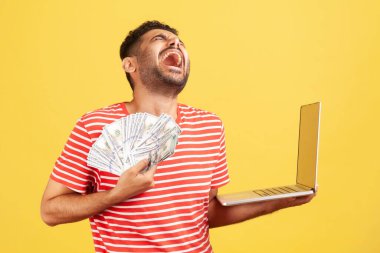 Çizgili tişörtlü, son derece mutlu bir adam elinde dizüstü bilgisayarla bağırıyor ve gülüyor ve çok para kazanıyor. Sarı arka planda kapalı stüdyo çekimleri