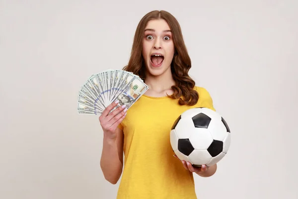 黄色のTシャツのサッカーボールを保持し 多くの紙幣で非常に興奮したティーンエイジャーの女の子は 喜んで叫んで カメラを見て 賭けと勝利 灰色の背景に隔離された屋内スタジオショット — ストック写真