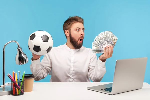 数百ドル札のバッチを見てショックを受けた興奮した男のオフィスの労働者サッカーボールを保持し ラップトップで職場に座って スポーツへの賭け 青い背景に隔離された屋内スタジオショット — ストック写真