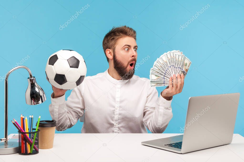 Shocked excited man office worker looking at batch of hundred dollar bills holding soccer ball, sitting at workplace with laptop, sports betting. Indoor studio shot isolated on blue background