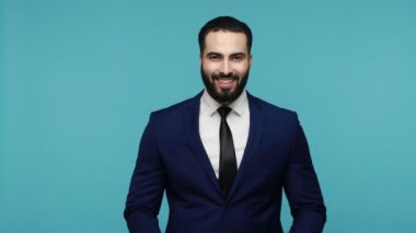 Siyah takım elbiseli, sakallı çekici bir adam, kameraya gülümseyerek bakıyor, başparmağını kaldırıyor, bir şeyler öneriyor, iyimserliğini dile getiriyor. Mavi arka planda kapalı stüdyo çekimi.