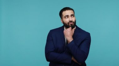 Siyah takım elbiseli, iş anlarını düşünen, doğru çözümü arayan, elini çenesinin altında tutan yakışıklı, düşünceli genç adam. Mavi arka planda kapalı stüdyo çekimi.