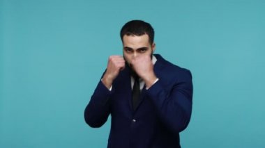 Siyah takım elbiseli ciddi bir genç adam savunma pozisyonunda duruyor ve yumruklarını dövüşmek için kaldırıyor, boks jesti yapıyor, antrenman yapıyor. Mavi arka planda kapalı stüdyo çekimi.