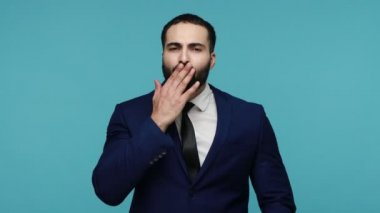 Seni seviyorum! Siyah takım elbiseli yakışıklı sakallı adam avuç içlerine hava öpücüğü yolluyor, flört ifadeleriyle kameraya bakıyor, romantizm gösterisi yapıyor. Mavi arka planda kapalı stüdyo çekimi.