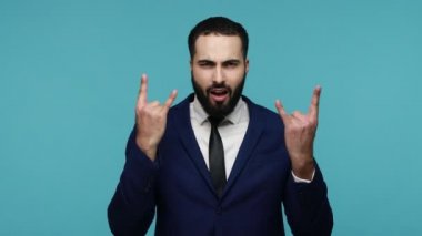 Resmi takım elbiseli, ateşli, çılgın esmer adam parmakları havada ve suratını buruşturarak, mutlu bir şekilde bağırarak rock and roll işareti gösteriyor. Mavi arka planda kapalı stüdyo çekimi.