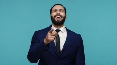 Çok komiksin! Takım elbiseli neşeli sakallı adam kamerayı gösterip gülüyor, heyecanla bakıyor, komik bir yüz ifadesi var. Mavi arka planda kapalı stüdyo çekimi.