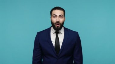 Resmi kıyafetli mutlu esmer adam gözlerine inanmadı, kameraya baktı zaferi ifade etti, yumruklarını sıktı, mutluluğu ifade etti. Mavi arka planda kapalı stüdyo çekimi.