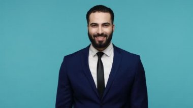 Hey sen, şanslı adam! Takım elbiseli neşeli, sakallı esmer adam kameraya heyecanla bakıyor piyango talihlisi, başarına seviniyor. Mavi arka planda kapalı stüdyo çekimi.