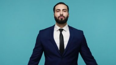 Siyah takım elbiseli genç sakallı iş adamı dikkatli yoga yapıyor, sakinleşmeye çalışıyor, meditasyon yapıyor, uyum ve dengeyi bulmaya çalışıyor. Mavi arka planda kapalı stüdyo çekimi.