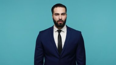 Bundan hoşlanmadım! Resmi takım elbiseli sakallı, tatmin olmamış yakışıklı bir adamın portresi baş parmakları aşağıda kameraya bakıyor. Mavi arka planda kapalı stüdyo çekimi.