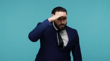 Siyah takım elbiseli özenli esmer adam dikkatlice uzaklara bakıyor, gözlerini dikmiş kamerayı işaret ediyor. Mavi arka planda kapalı stüdyo çekimi.