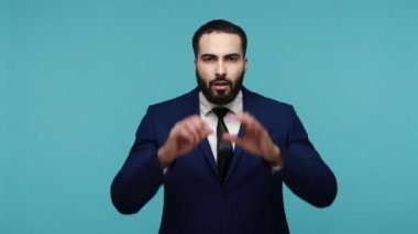 Resmi takım elbiseli kızgın esmer sakallı adam ellerini açık tutuyor ve her yandan bağırıyor, yüksek sesle bağırıyor, dikkat ediyor. Mavi arka planda kapalı stüdyo çekimi.