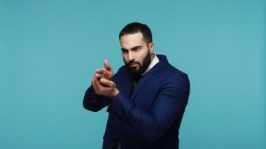 Siyah takım elbiseli sakallı adam kovboy gibi davranıyor, parmak tabancasını kavrayıp kamerayı doğrultuyor, el kol hareketi yapıyor, öldürüyor. Mavi arka planda kapalı stüdyo çekimi.