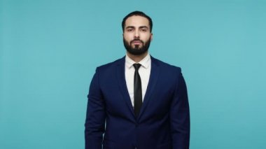 Resmi tarzda giyinmiş sakallı bir adam el sallıyor, evet diyor ve kameraya olumlu bir ifadeyle bakıyor, düzeni dinliyormuş gibi yapıyor. Mavi arka planda kapalı stüdyo çekimi.