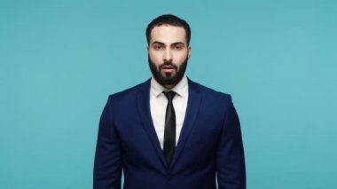 Hoşnutsuz esmer, resmi takım elbiseli, sakallı bir adam parmağıyla aptalca bir jest yapıyor. Üzücü ve bunalımlı bir ifade takınıyor. Mavi arka planda kapalı stüdyo çekimi.