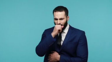 Sağlıksız genç iş adamı siyah resmi takım elbise giyip öksürerek ağzını eliyle kapatıyor, hasta hissediyor, grip belirtileri gösteriyor, zatürree olma riski var. Mavi arka planda kapalı stüdyo çekimi.