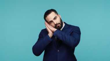 Resmi tarz takım elbise içinde yorgun, uykulu genç bir iş. Uykusu geliyor, uyukluyor, avuçlarını kulaklarının yanında tutuyor, gözlerini kapatıyor. Mavi arka planda kapalı stüdyo çekimi.