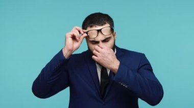 Resmi stil kıyafetli yorgun işadamı parmaklarıyla ovuşturuyor, gözlük altında yorgun gözlerle uzun saatler çalışıyor, çok çalışıyor. Mavi arka planda kapalı stüdyo çekimi.