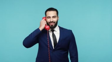 Çağrı merkezi konsepti. İş elbisesi giymiş pozitif sakallı bir adam bana jest yaparken ve telefonla konuşurken kameraya gülümsüyor. Mavi arka planda kapalı stüdyo çekimi.