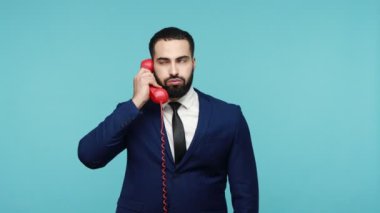 Resmi tarzda giyinmiş kızgın, sakallı genç bir adam elinde kırmızı bir telefonla bağırıyor, biriyle anlaşmazlık yaşıyor. Mavi arka planda kapalı stüdyo çekimi.