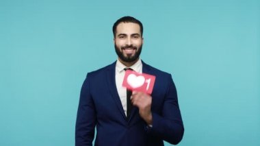 Yakışıklı sakallı blogcu takım elbiseli adam mutlu bir gülümsemeyle kameraya bakıyor, sosyal medyayı simge ve başparmaklar gibi gösteriyor, kalp düğmesine basıyor. Mavi arka planda kapalı stüdyo çekimi.