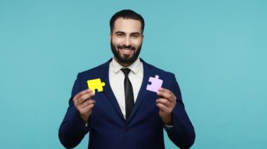 Takım elbiseli çekici sakallı iş adamı elinde iki parça yapboz tutuyor, renkli yapboz, başarılı bir iş birliğinden memnun, görevleri çözmekten. Mavi arka planda kapalı stüdyo çekimi.