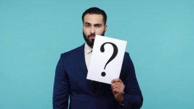 Kafası karışmış sakallı işadamı elinde soru işareti olan bir kağıt tutuyor, problem çözümü arıyor, şüpheli bir ifadeyle fikir üretiyor. Mavi arka planda kapalı stüdyo çekimi.