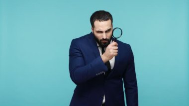 Resmi stil takım elbiseli çekici, koyu renk saçlı bir dedektif elinde büyüteçle kameraya bakıyor. Mavi arka planda kapalı stüdyo çekimi.
