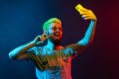 Başarılı, heyecanlı sakallı hippi blog yazarı, yeşil saçlı, modaya uygun kot tulumlu, Ziyaretçi jesti yapan, telefon kamerasına poz veren, selfie çeken. Renkli neon ışık, kapalı stüdyo çekimi..