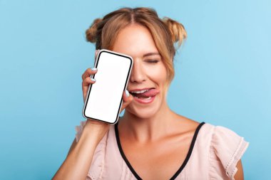 İki komik saçlı, güzel sarışın kadının portresi. Yüzünün yarısını telefonla kapatmış, gözlerini kapatmış, dilini göstermiş. Mavi arka planda kapalı stüdyo çekimi.