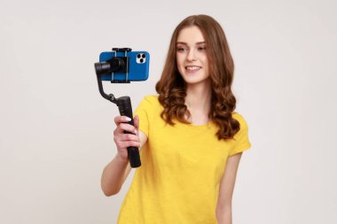 Tişörtlü, genç ve modaya uygun güzel bir kadın kameralı akıllı telefonu elinde tutuyor, blogcu sosyal ağ için selfie çekiyor, canlı yayında. Gri arka planda izole edilmiş kapalı stüdyo görüntüleri.