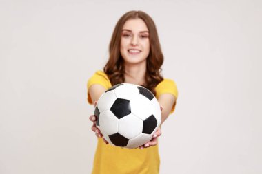 Genç yaşta güzel kahverengi saçlı bir bayan kameraya bakıyor, hoş bir gülümsemeyle futbol topu sunuyor, futbol maçı oynamak istiyor. Gri arka planda izole edilmiş kapalı stüdyo görüntüleri.