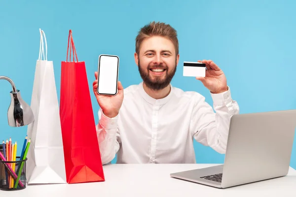 スマートフォンやクレジットカードを持っている積極的な笑顔の男は オンラインで買い物をし ノートパソコンやショッピングバッグで職場に座ってキャッシュバックを受け取ります 青い背景に隔離された屋内スタジオショット — ストック写真