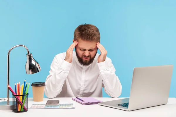 疲れ切った人事務員は 机の上のノートパソコンで職場に座って耐えられない頭痛を感じる寺院をマッサージし 片頭痛に苦しむハードタスクを解決します 青い背景に隔離された屋内スタジオショット — ストック写真