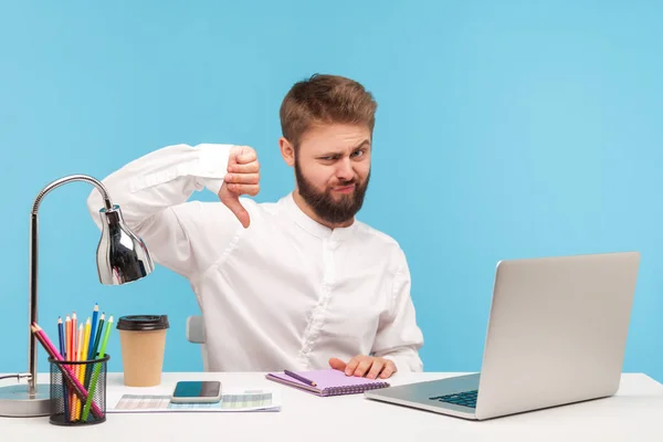 Dislike! Displeased man employee sitting office workplace showing thumbs down gesture, expressing disapproval, negative service rating. Indoor studio shot isolated on blue background