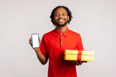 Rastgele giyinmiş kırmızı tişörtlü gülümseyen bir adam, elinde online alışveriş reklamları için boş ekranlı bir hediye kutusu ve cep telefonu tutuyor. Gri arka planda izole edilmiş kapalı stüdyo görüntüleri.
