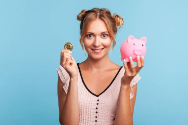 İki komik saçlı mutlu sarışın kadın, kameraya altın kripto sikke ve kumbara gösteriyor, bitcoin ve kripto para yatırıyor. Mavi arka planda kapalı stüdyo çekimi.