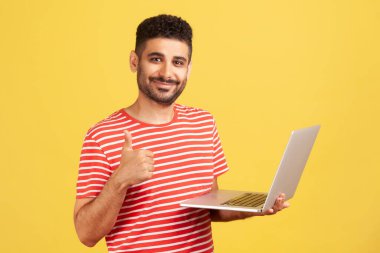 Çizgili tişörtlü, sakallı, elinde dizüstü bilgisayarla tele çalışmaktan memnun bir şekilde başparmak kaldıran pozitif gülümseyen serbest bir adam işini seviyor. Sarı arka planda kapalı stüdyo çekimleri