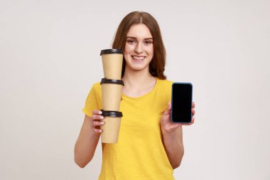 Taşınabilir uygulama, internet reklamı. Sarı tişörtlü mutlu genç kız elinde kahve fincanları ve cep telefonuyla sergi düzenliyor. Gri arka planda izole edilmiş kapalı stüdyo görüntüleri.