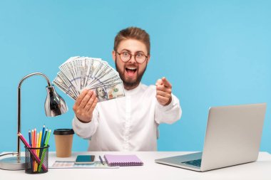 Son derece pozitif sakallı zengin bir adam elinde dolarlarla seni işaret ediyor, iş yerinde dizüstü bilgisayarla oturuyor, seni büyük para kazanmaya motive ediyor. Mavi arka planda kapalı stüdyo çekimi