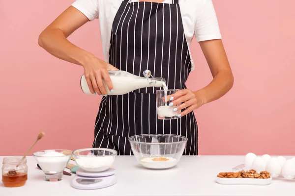 Cropped image of female confectioner or baker making dough for cakes or cookies, pours milk to glass for adding to flour, homemade pastry. Indoor studio shot isolated on pink background.